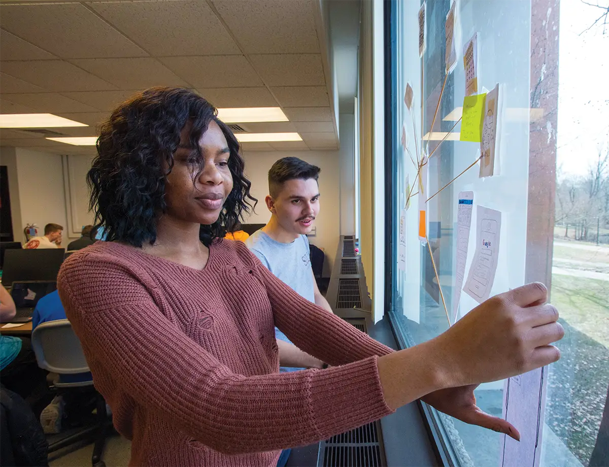 Two students review wireframe sketches and sticky notes arranged on a classroom window during a design project critique.