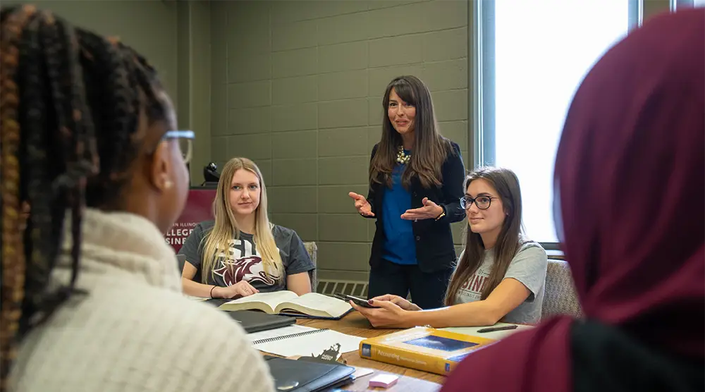 **Alt text:** A faculty member stands at a table speaking with a small group of students in a seminar room, with an SIU College of Business banner visible in the background.