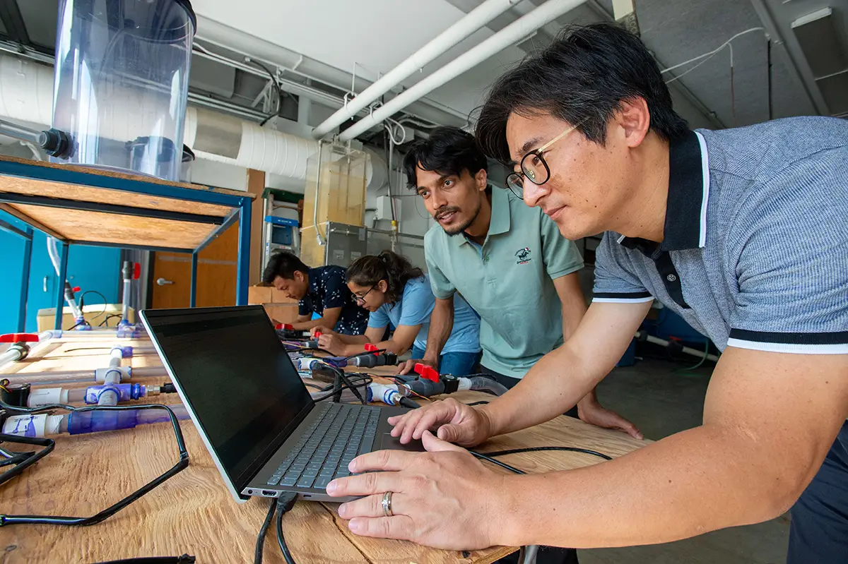 A faculty member and students lean over a workbench in an engineering lab, reviewing data on a laptop surrounded by pipes, sensors, and research equipment.