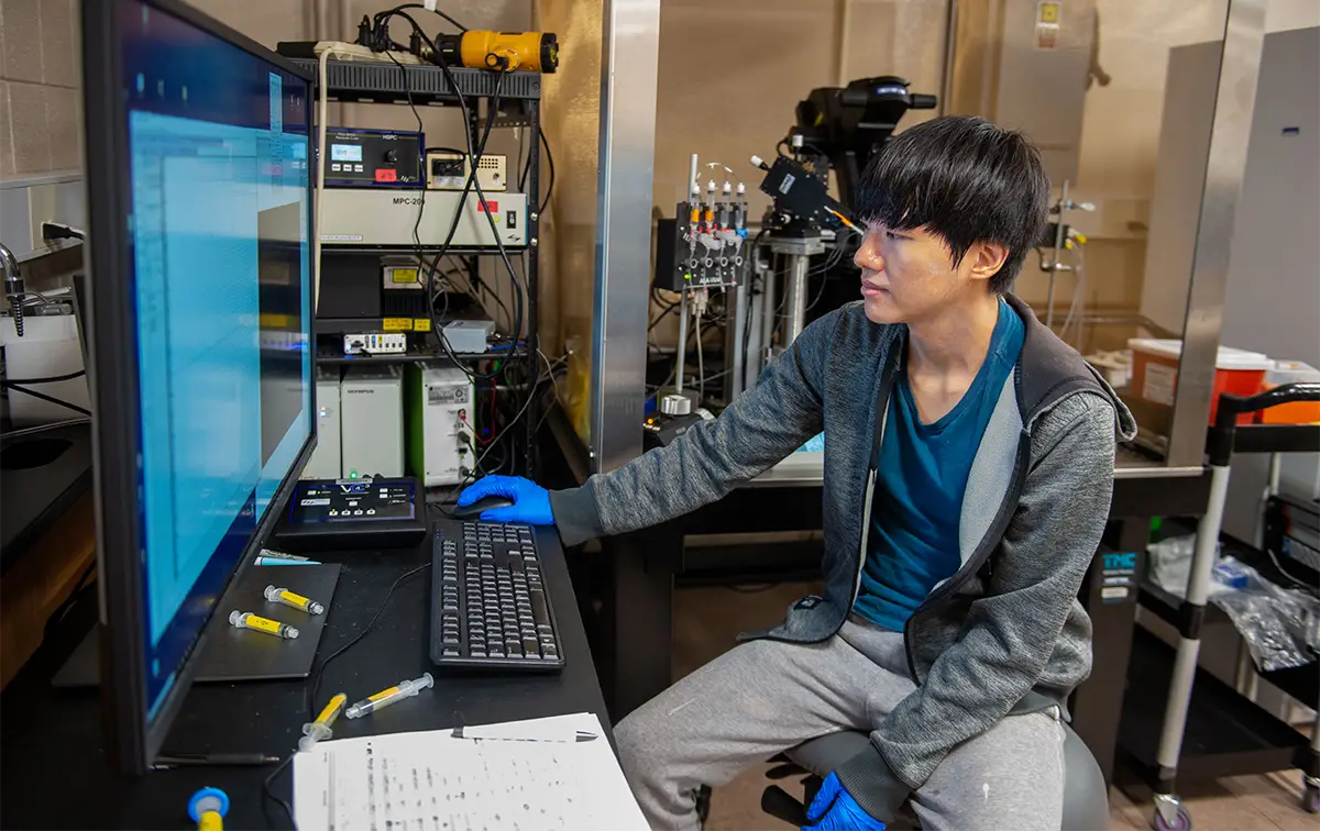 A student wearing blue gloves sits at a workstation in a research laboratory, operating a computer surrounded by scientific equipment and instruments.