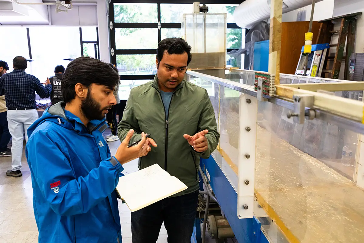 Two students discuss observations next to a water channel flume in an engineering laboratory, with one holding a notebook and other students visible in the background.