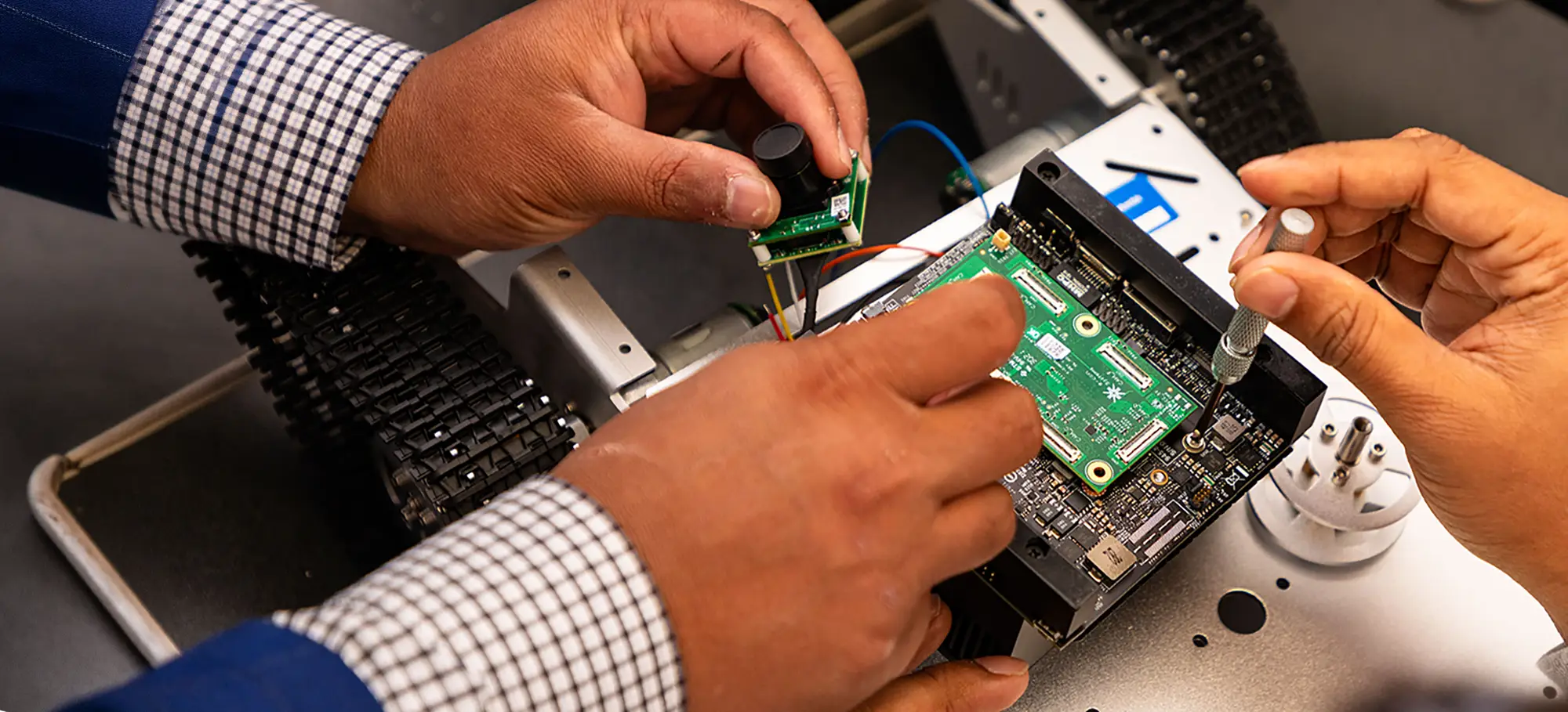 Two people install a circuit board and camera module into a tracked robotic vehicle in an electronics lab.