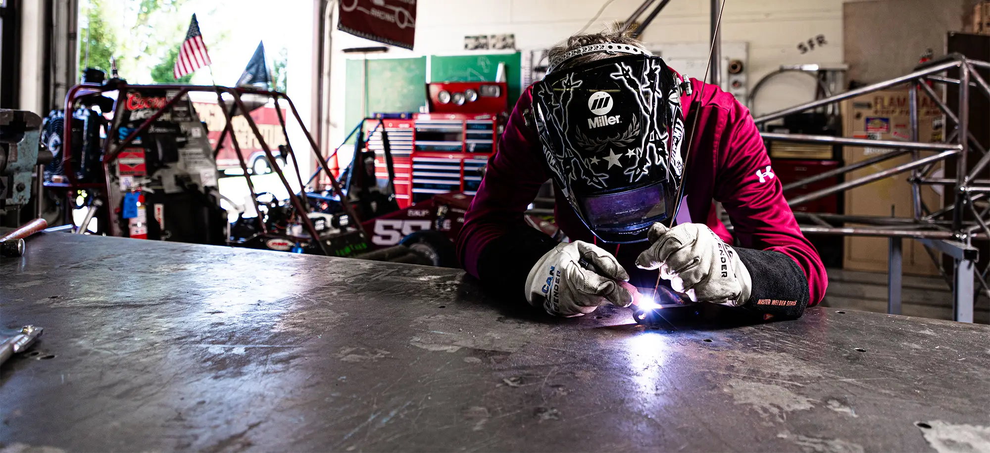 A student wearing a Miller welding helmet and protective gloves welds a metal surface in an automotive engineering shop, with a race car frame visible in the background.