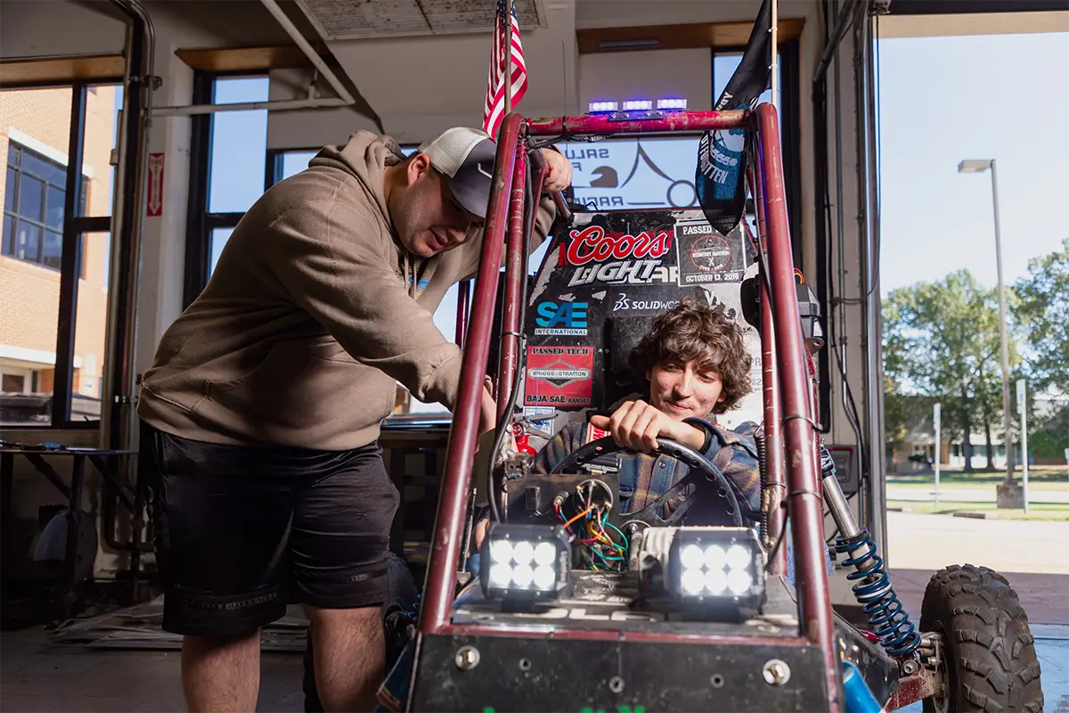 Two students work on an off-road vehicle outside an engineering shop, with one seated in the cockpit and the other making adjustments to the roll cage.