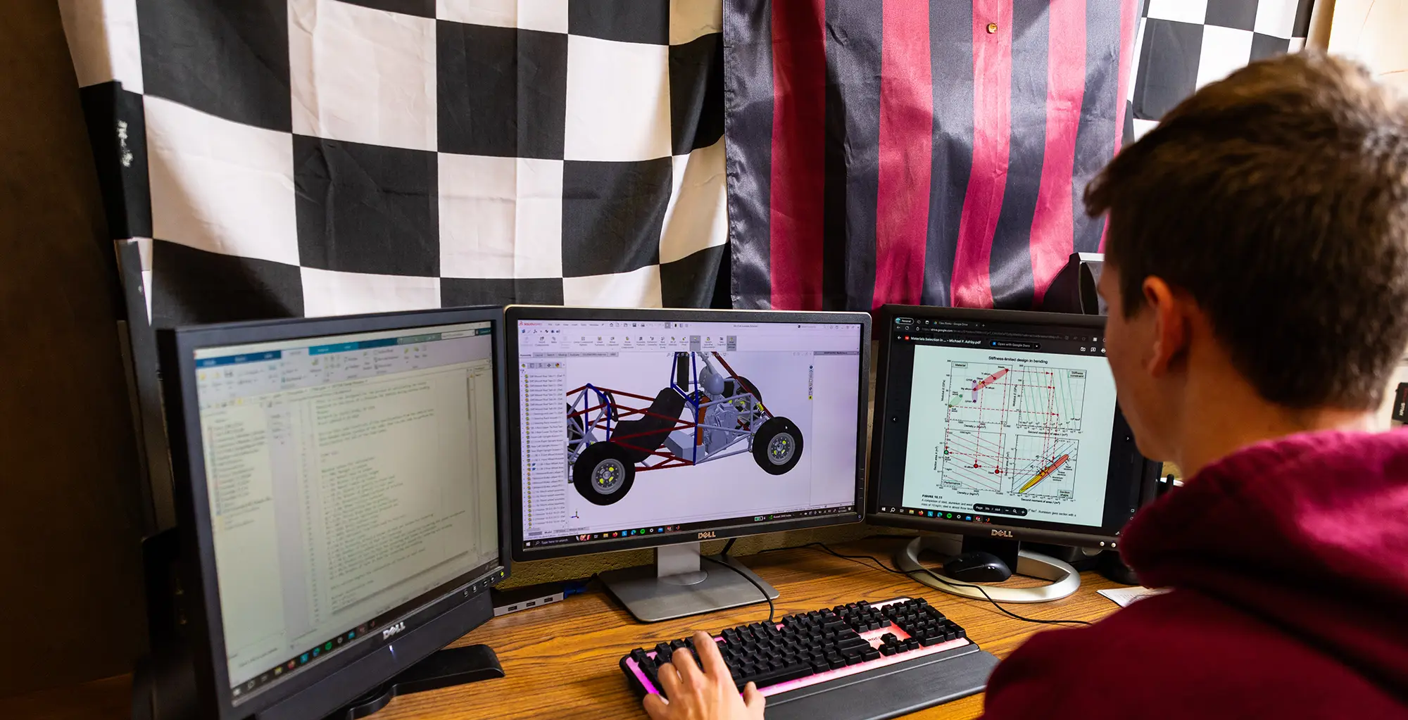 A student works at a three-monitor workstation reviewing a CAD model of a race car frame alongside engineering diagrams, with a checkered flag displayed on the wall behind them.
