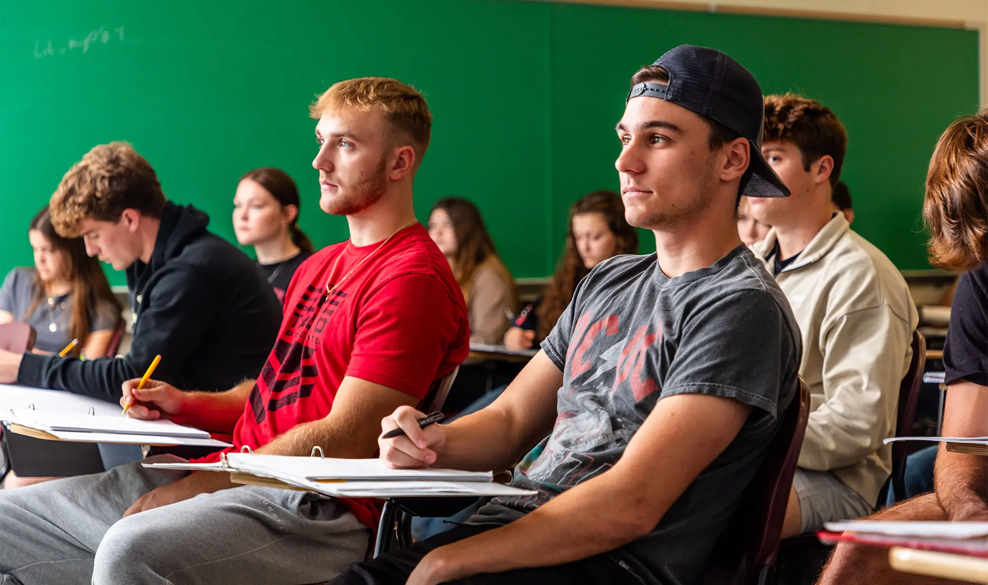 Students sit at desks with notebooks in a college classroom, with a green chalkboard visible in the background.