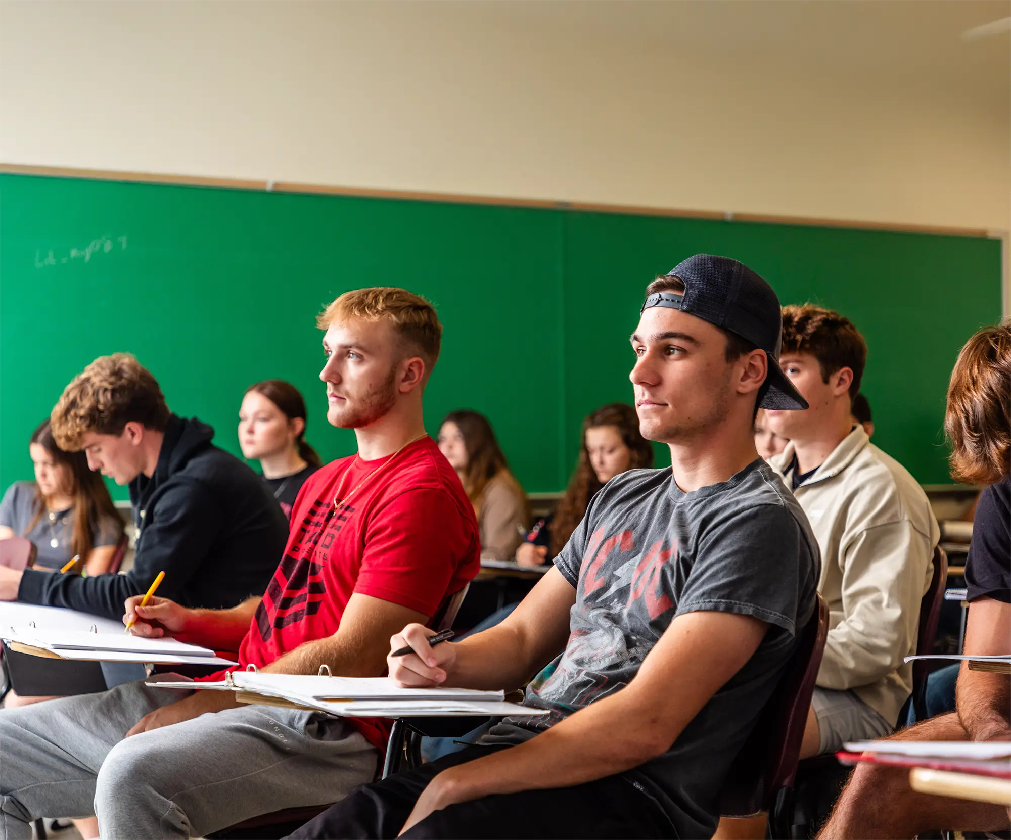 Students sit at desks with notebooks in a college classroom, with a green chalkboard visible in the background.