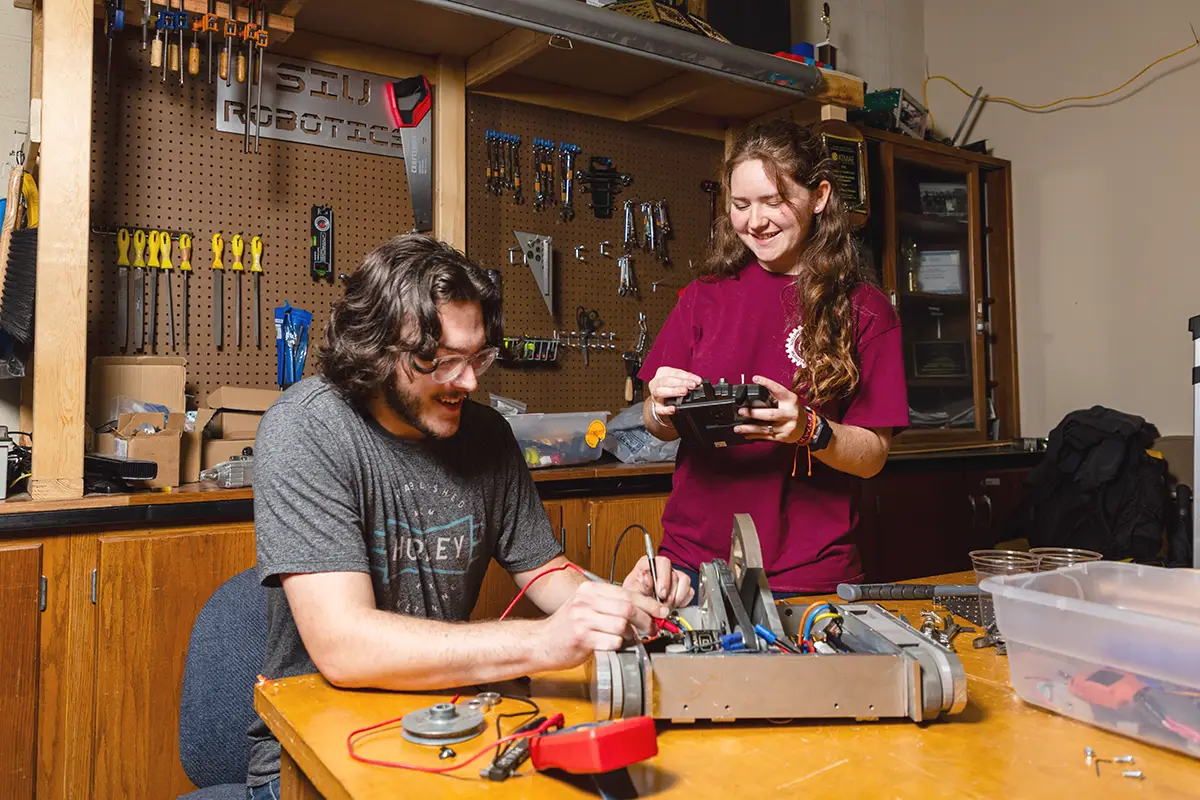 Two students work on a robot in the SIU Robotics workshop, with one making adjustments to the machine and the other holding a remote control.