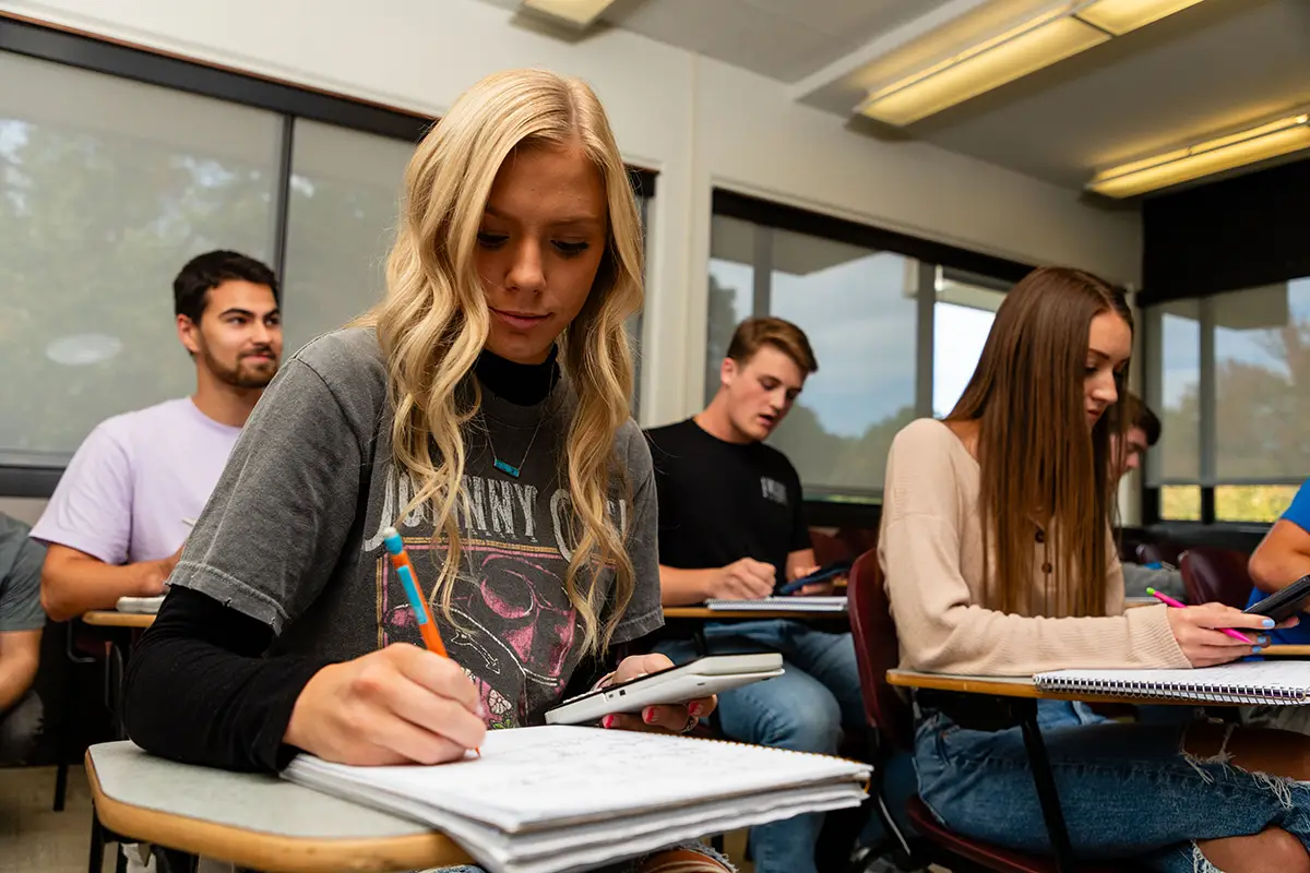 Students take notes at their desks during a college classroom lecture.
