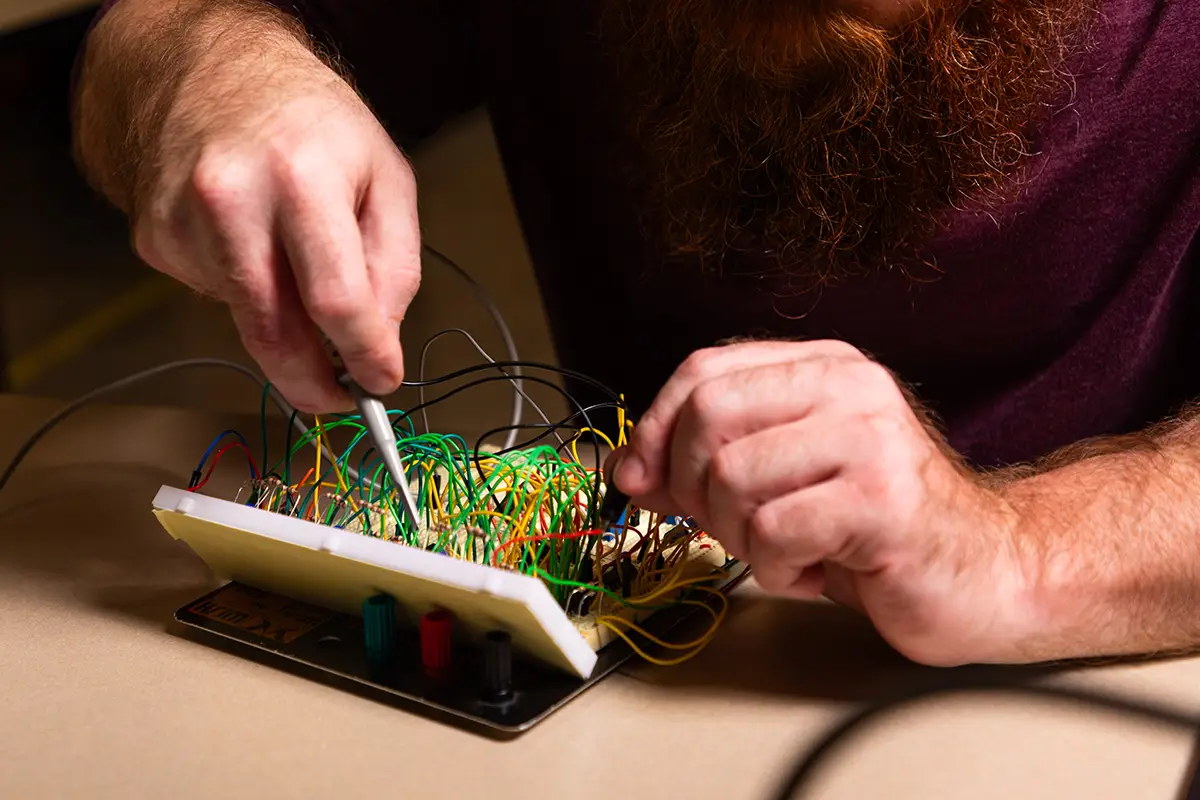 A student works with a probe on a densely wired breadboard circuit in an electronics lab.