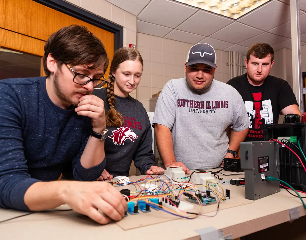 Four SIU students work together on an electronics wiring project at a lab bench, with circuit components and wiring spread across the table.