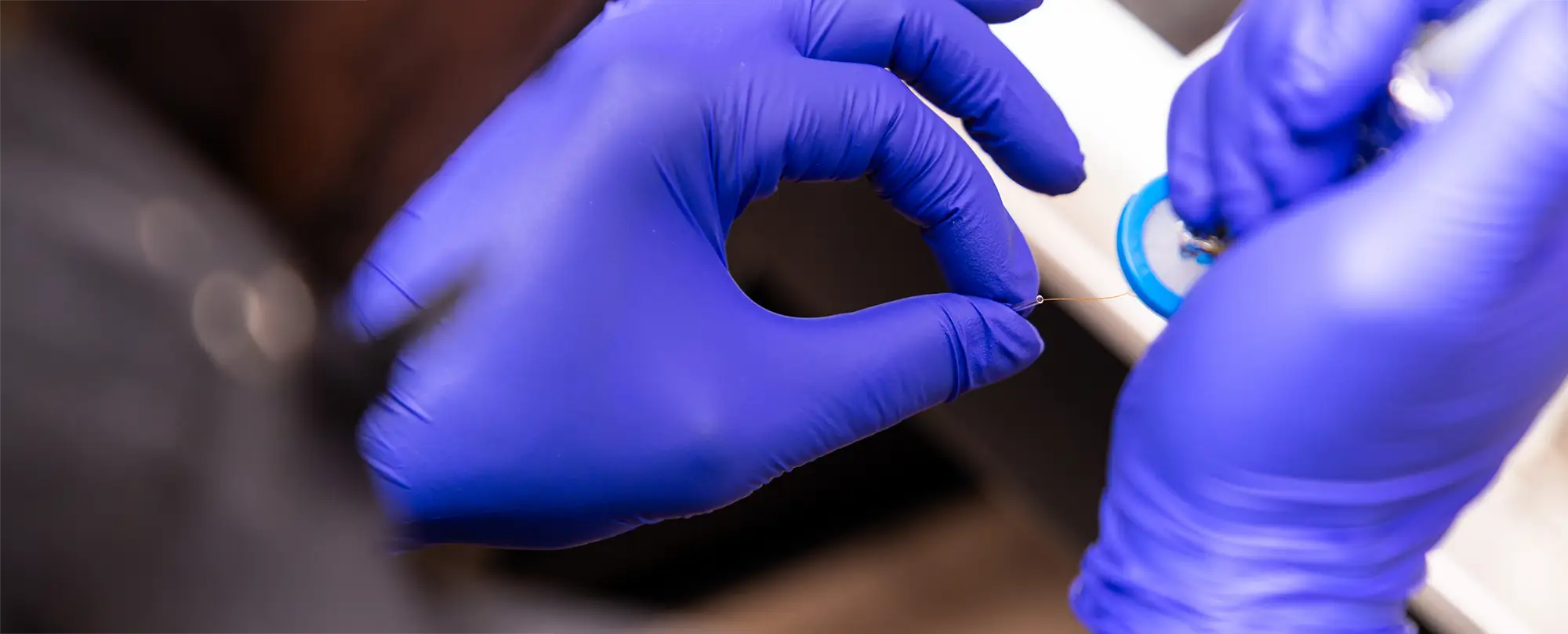 A close-up of gloved hands carefully handling a fine wire or filament in a laboratory setting.