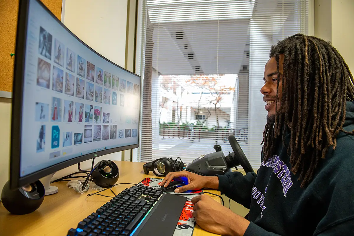 A student works at a curved ultrawide monitor displaying a grid of digital images, with headphones and a VR headset on the desk nearby.
