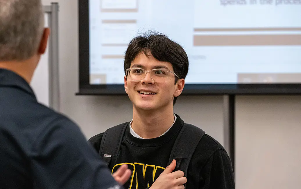 **Alt text:** A student wearing glasses and a backpack talks with an instructor in a classroom, with a projected presentation visible on a screen in the background.
