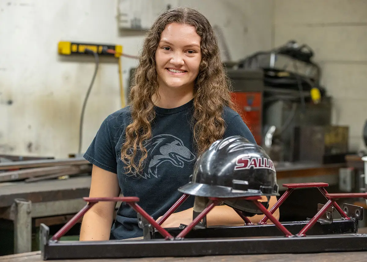 A student in an SIU t-shirt sits at a workbench in an engineering shop with a small steel bridge model and a Saluki hard hat in front of her.