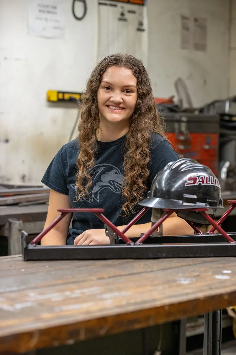 A student in an SIU t-shirt sits at a workbench in an engineering shop with a small steel bridge model and a Saluki hard hat in front of her.