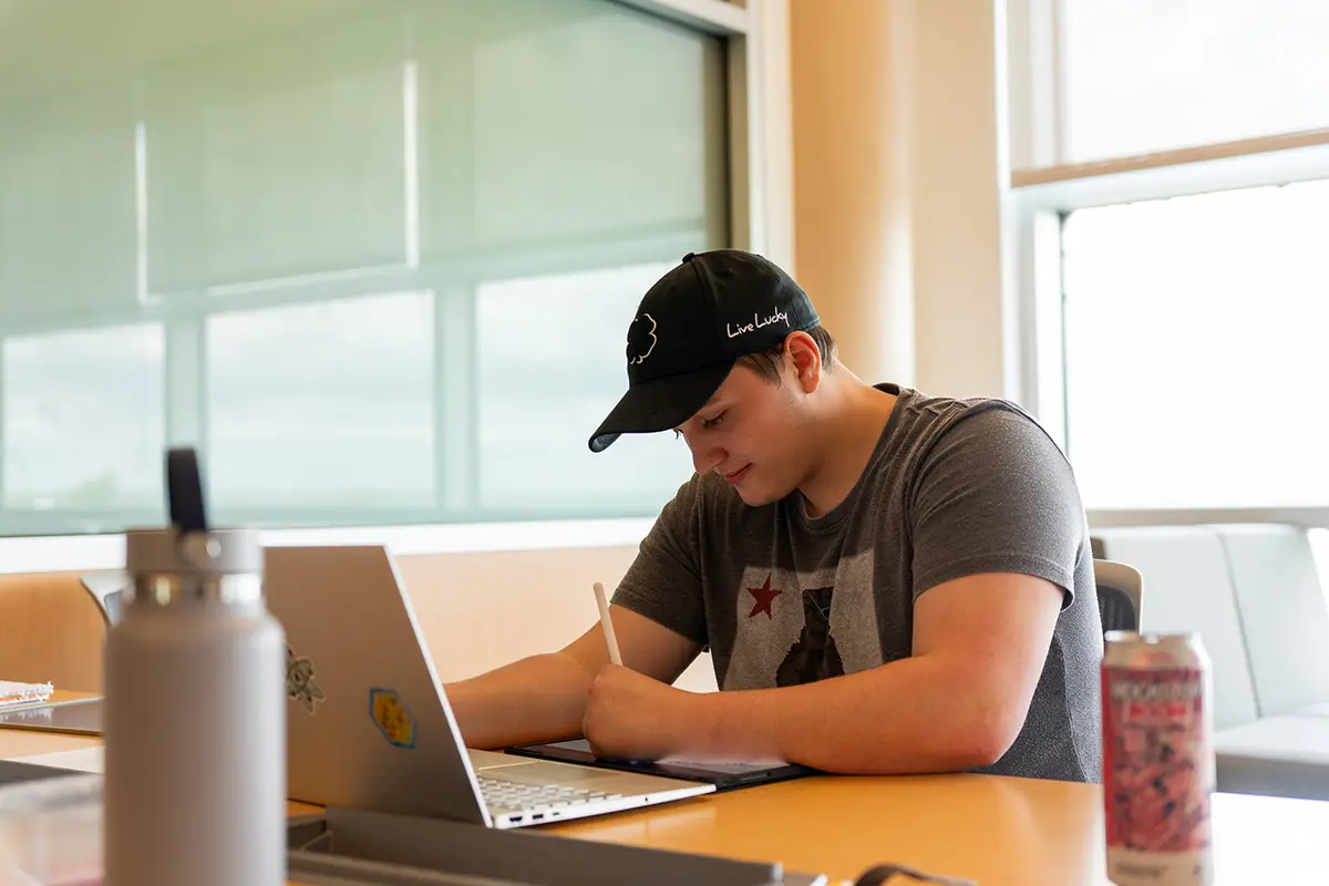 A student writes notes at a table with a laptop and drawing tablet in a bright campus study space.