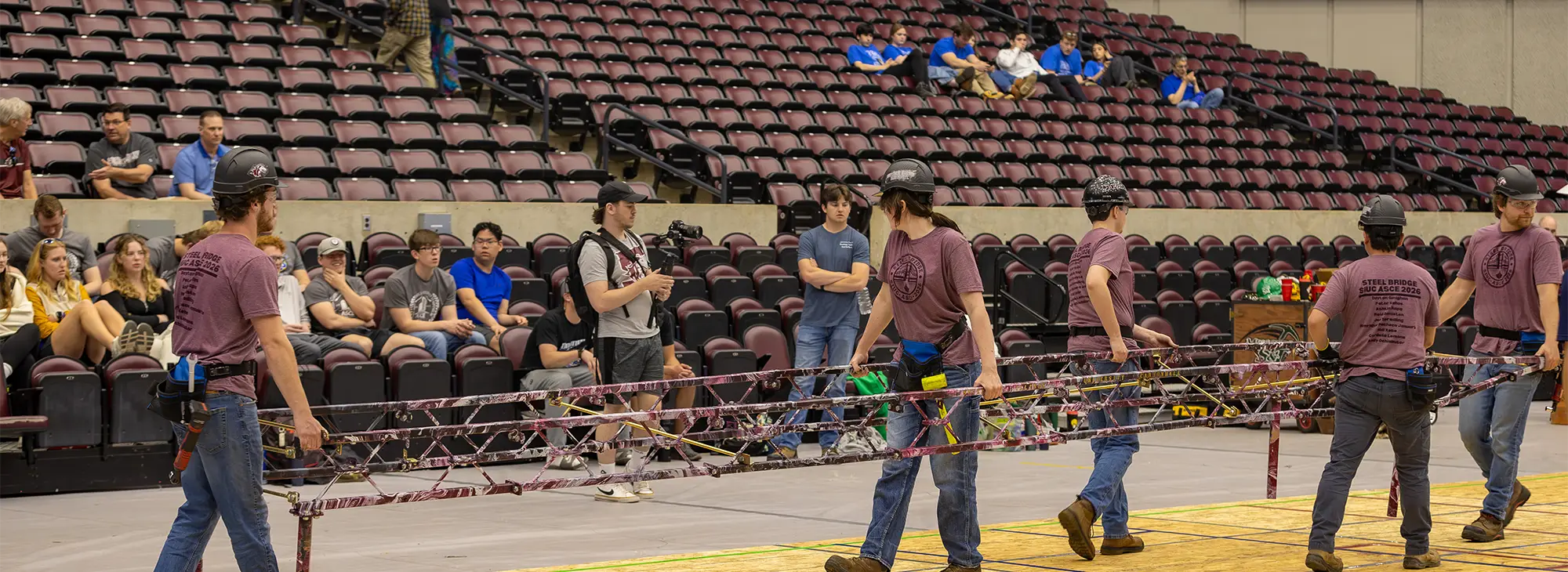 A team of students wearing hard hats and matching Steel Bridge competition shirts carry a steel truss structure across an arena floor during a collegiate engineering competition.