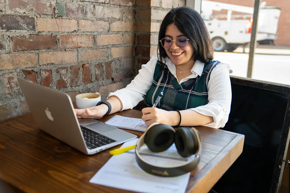 A student takes notes while working on a laptop at a table near a brick wall and window.