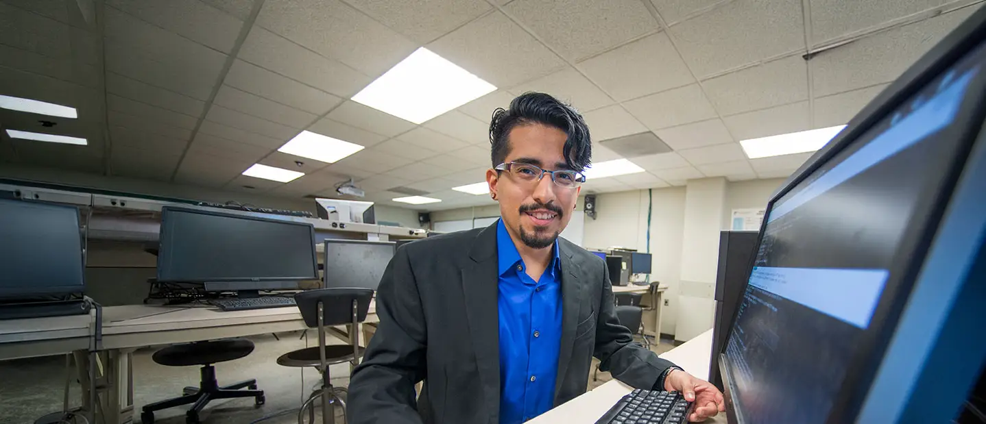 A student sits at a desktop computer in a university computer lab, with monitor.